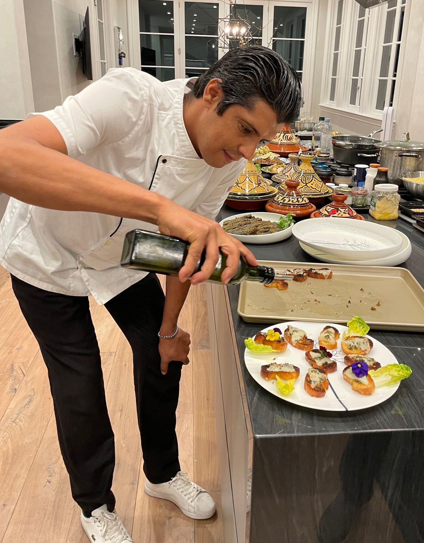 Chef Hicham Foual in chef's whites, finishing a tray of canapés with olive oil, clay tagines lined up behind him.