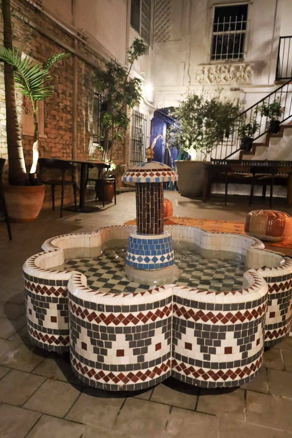 The courtyard at night: a zellige-tiled fountain in green, white and red, ringed by potted palms and lantern light.