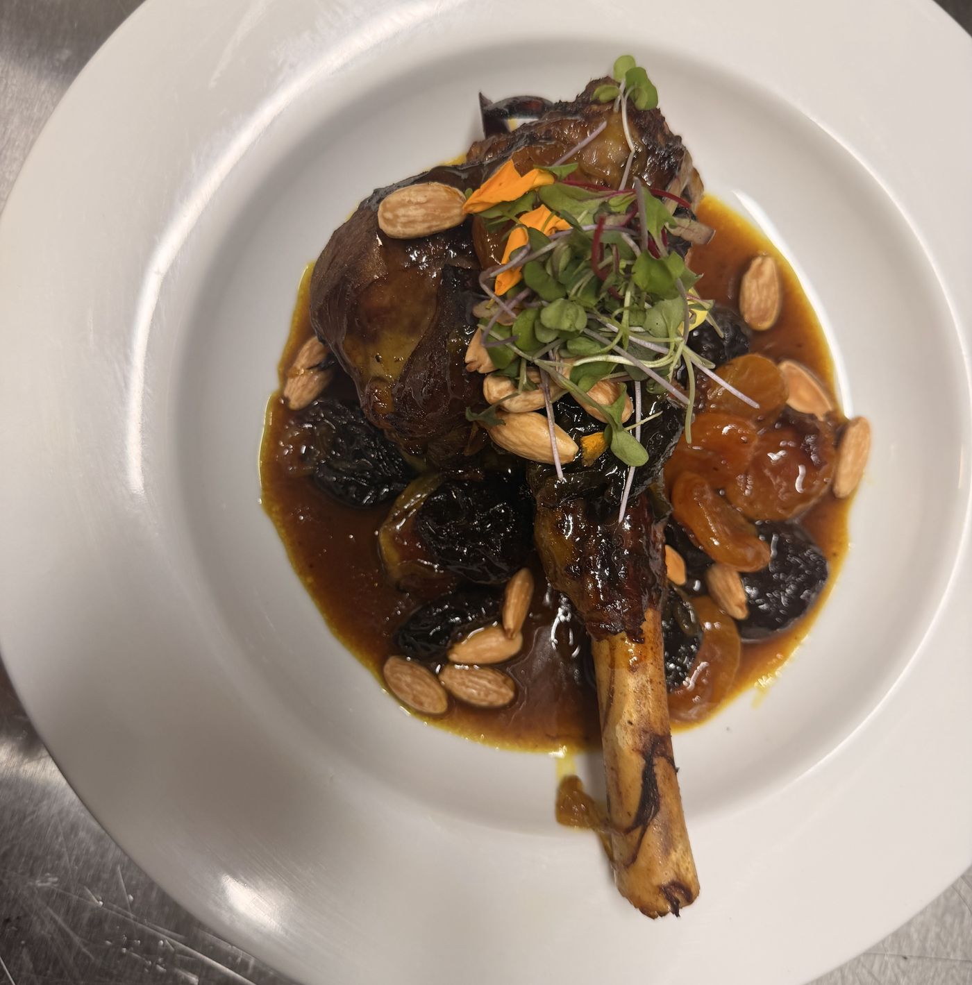 Lamb shank glazed dark and glossy, scattered with toasted almonds and prunes, on a white plate.