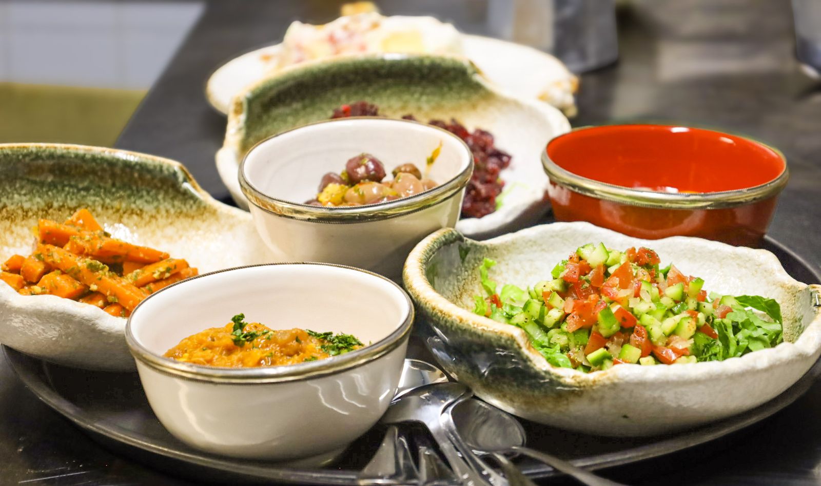 Mezze set out on the pass before service: carrots, beets, eggplant and a chopped tomato salad in white and green bowls.