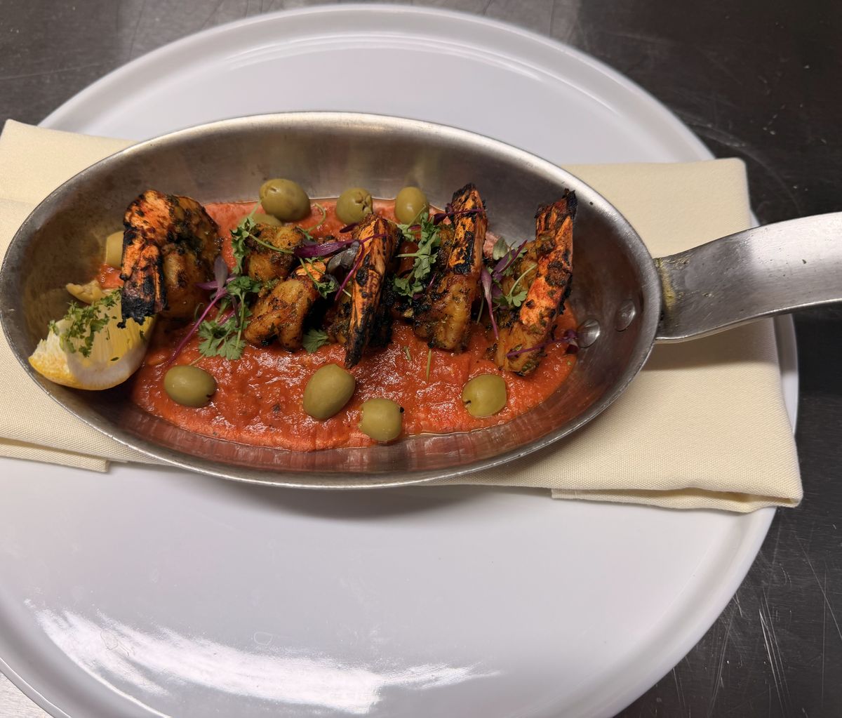 Grilled shrimp in a spiced tomato sauce with green olives and lemon, served in a steel pan.
